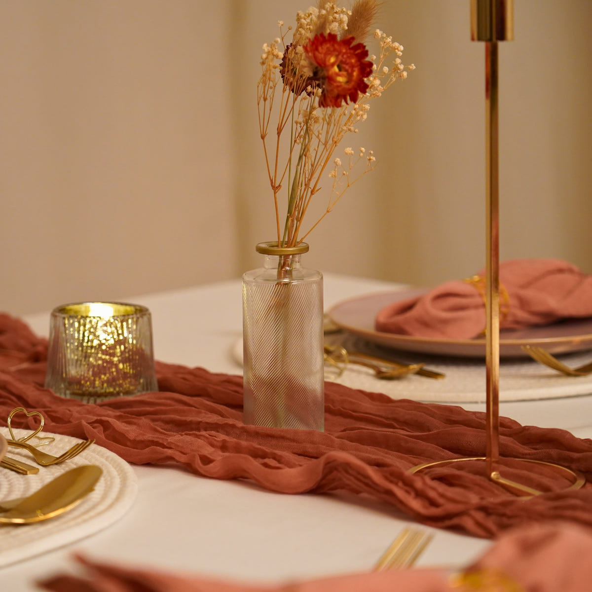 Trockenblumen in einer Glasvase mit Goldrand auf einem Tisch mit terracottafarbenem Läufer und goldenem Kerzenständer